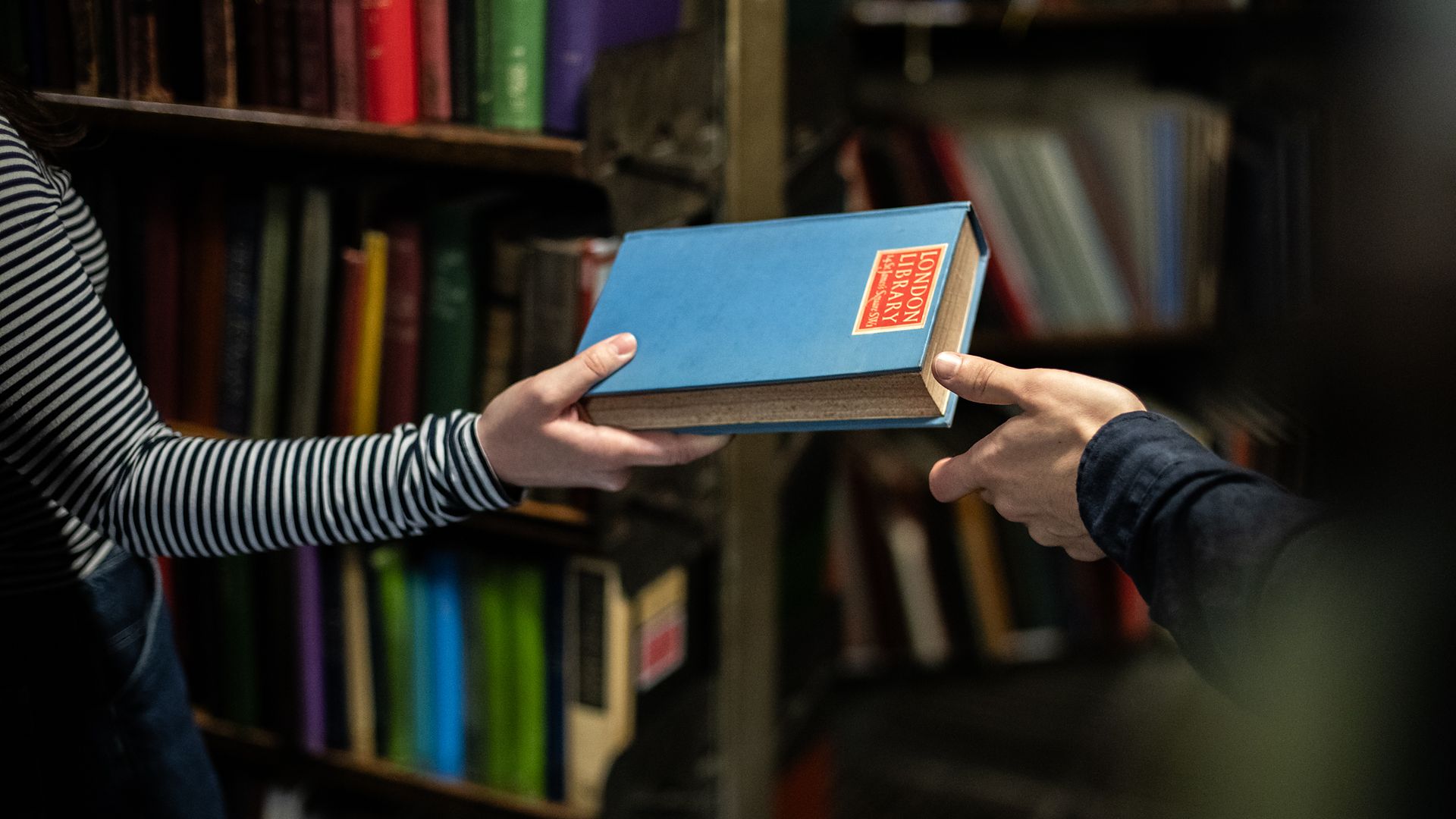One member passing a blue library book with a red London Library label to another.