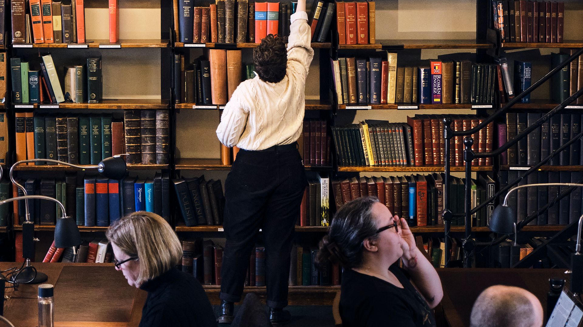 A member reaching for a volume on a high shelf as readers work at desks in the foreground.