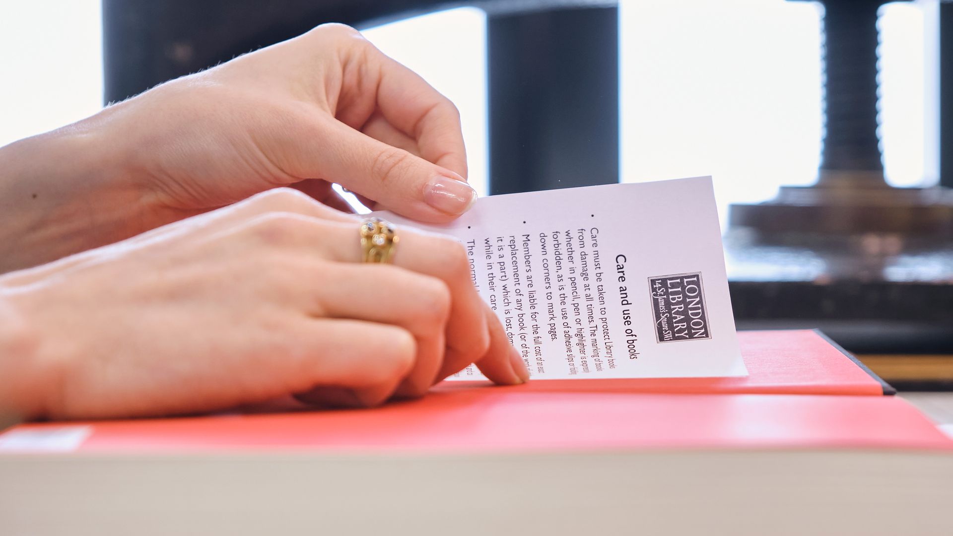 Hands tucking a care-and-use leaflet into a pink-bound book beside a press.