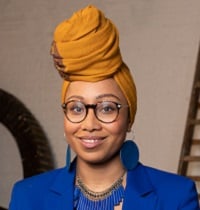 A portrait of Yassmin, a black woman with a yellow headscarf, blue jacket and glasses. 