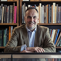 Portrait of a bearded white man in a blue shirt tan jacket in front of bookshelves. 