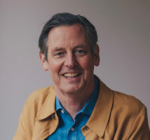 A portrait of Charles, a white man with dark hair in a yellow jacket and blue shirt.