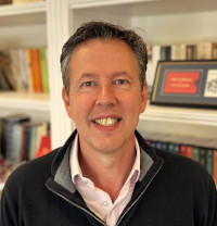 Portrait of Steven, a white man with dark hair in a pink shirt and pullover. 