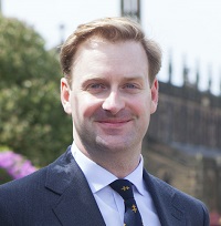 Portrait of Stephen, a white man with blonde hair in a grey suit and tie. 