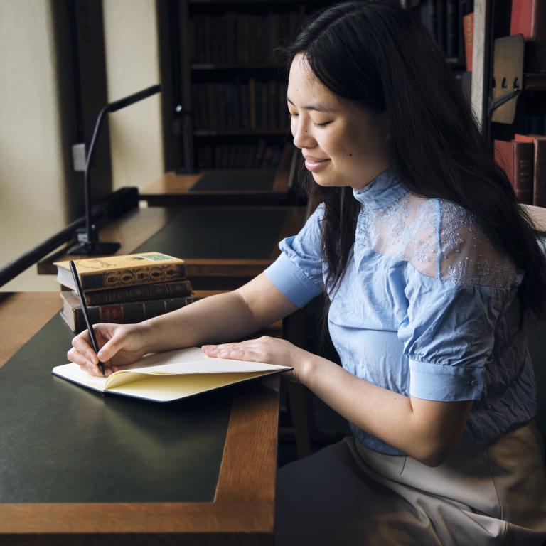 woman writing in a notebook at a desk in the Library