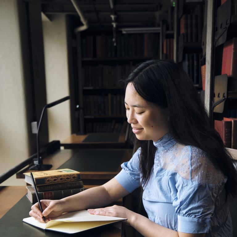 Woman writing in notebook with stack of books at desk