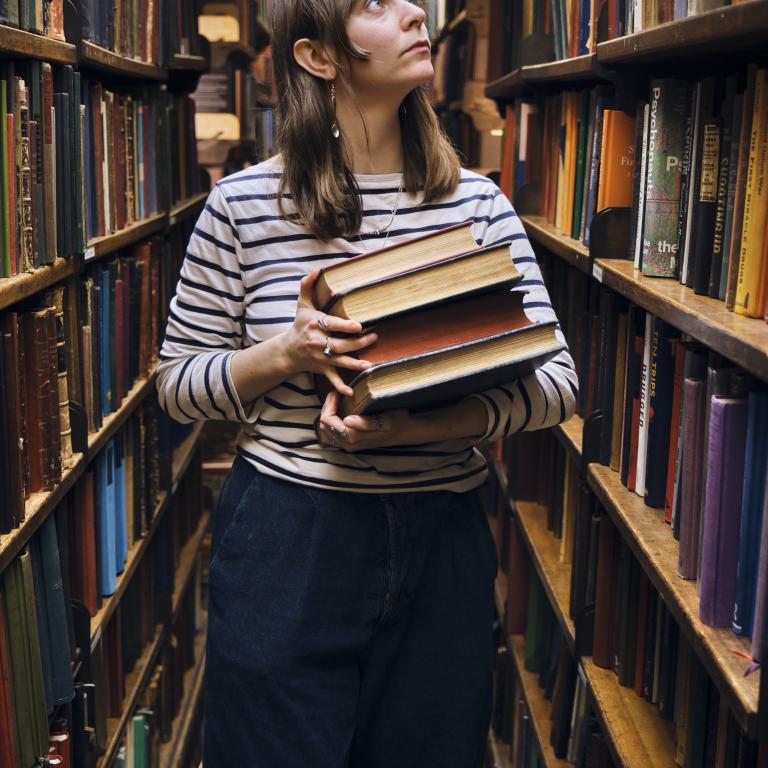 Woman walking through shelves at the library holding a stack of books