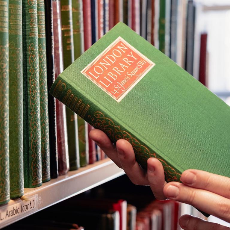 A hand taking a green cloth-bound book with a red London Library label from the shelf.