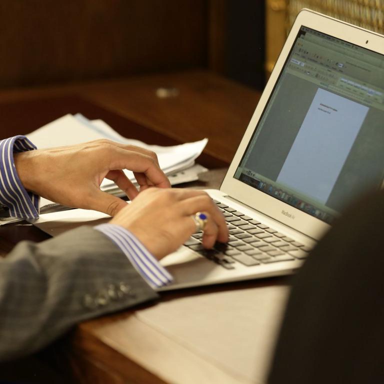 A close view of hands typing on a laptop at a reading desk, shirt cuffs showing.