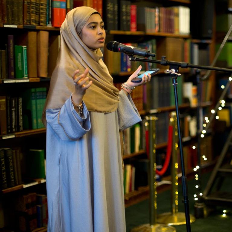 A performer reads at a microphone in front of the bookshelves at the Folio Prize event.