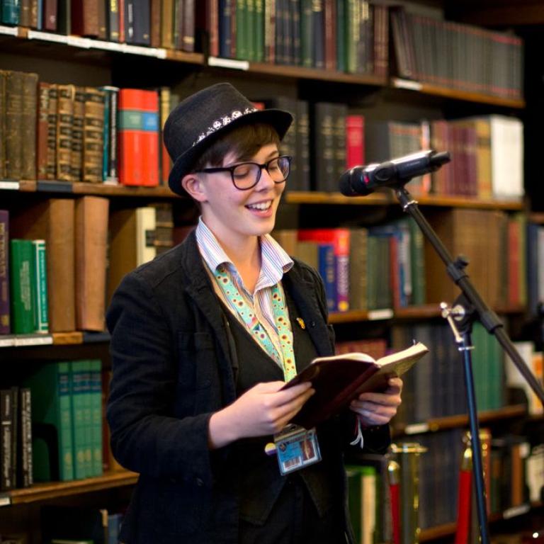 A performer in a bowler hat reads from a book at a microphone among the bookstacks.