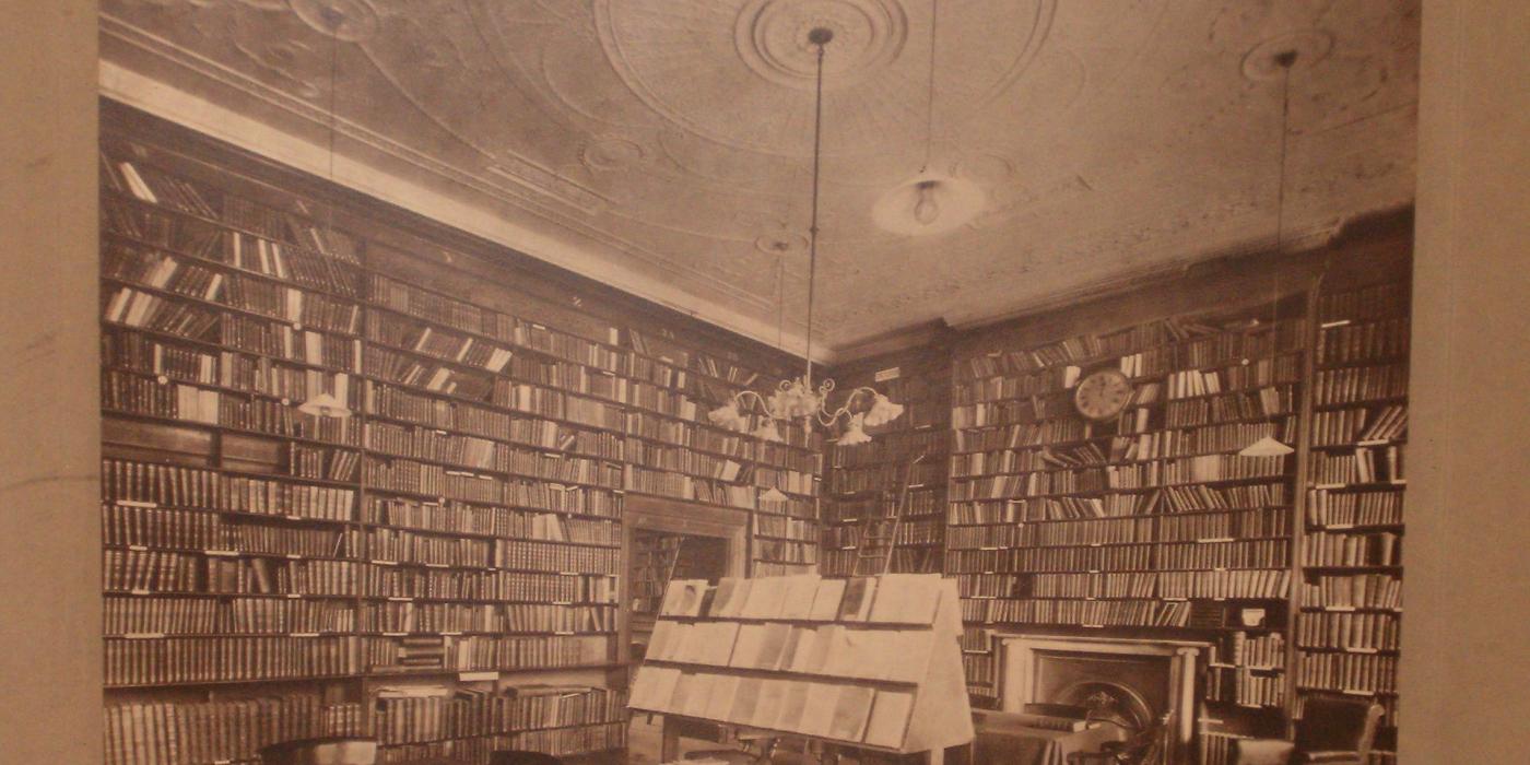 A sepia photograph of an 1880s reading room, its walls lined with books beside a fireplace.