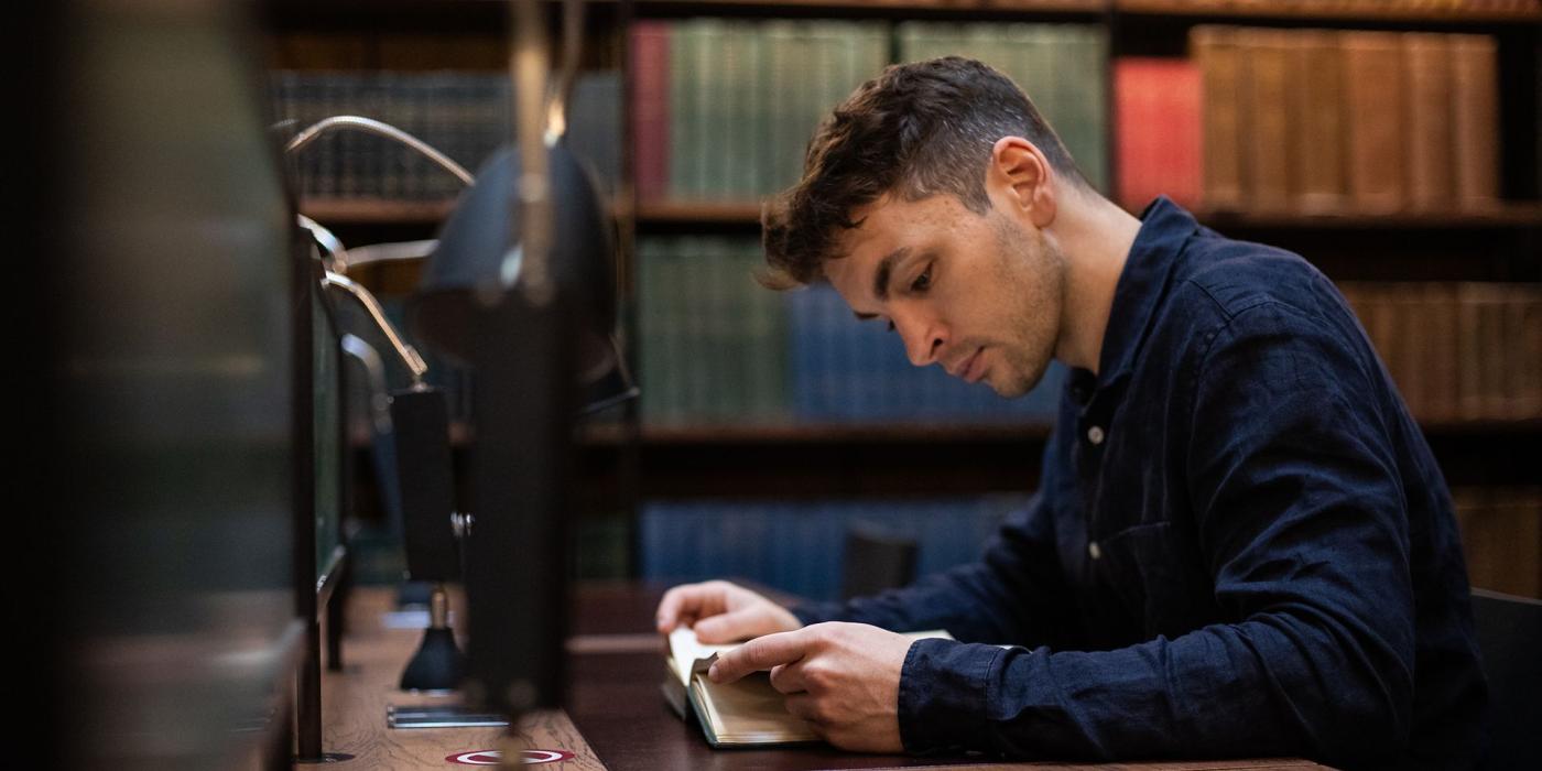 A member reading at a lamp-lit desk in front of shelves of bound volumes.