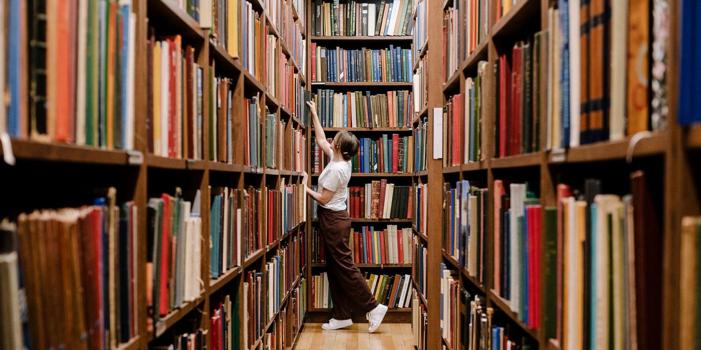 A member reaching up to a high shelf in a long, book-lined stack aisle.