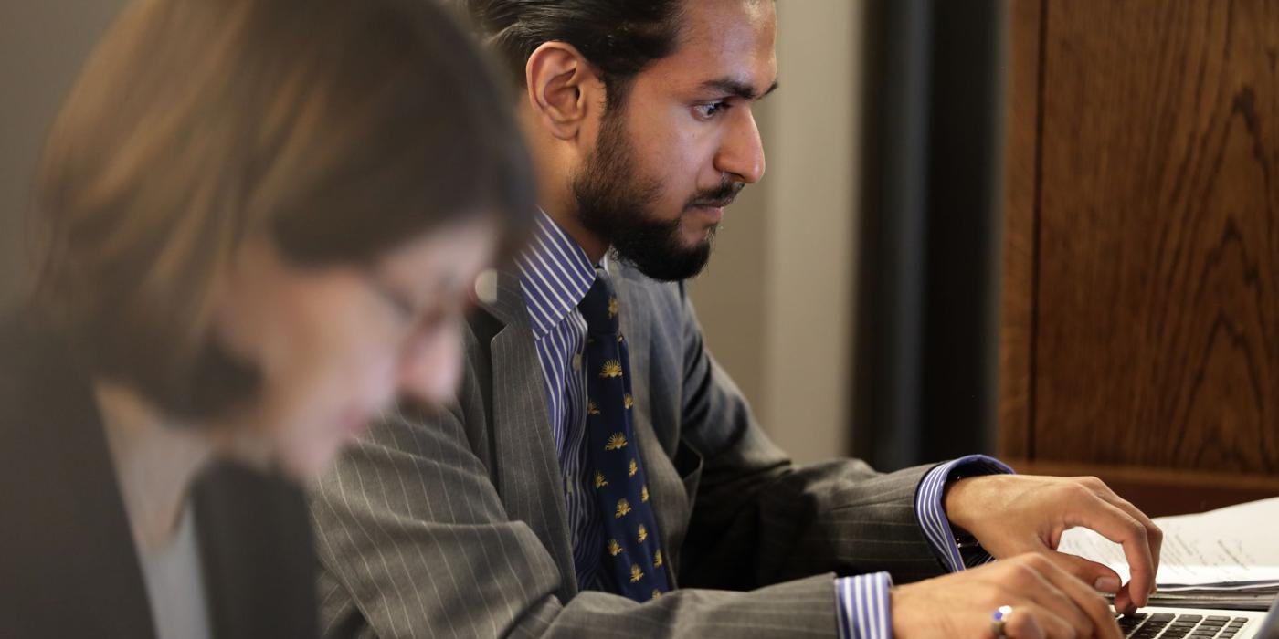 A member in a suit concentrating at a laptop, another reading in the foreground.