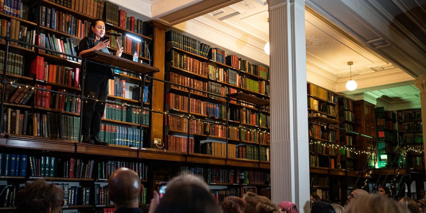 A poet performs from a lectern on the gallery to an audience in the Reading Room.