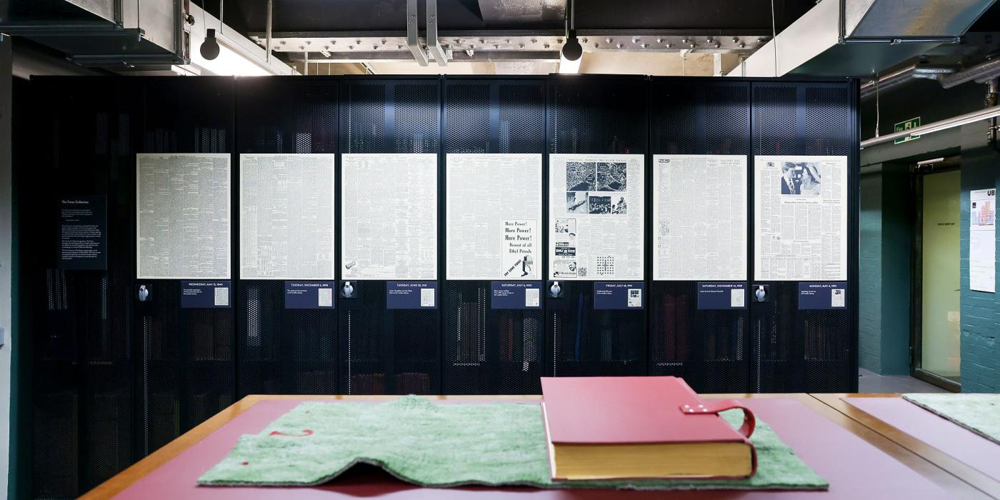 A large red bound newspaper volume on green baize, framed historic front pages behind.