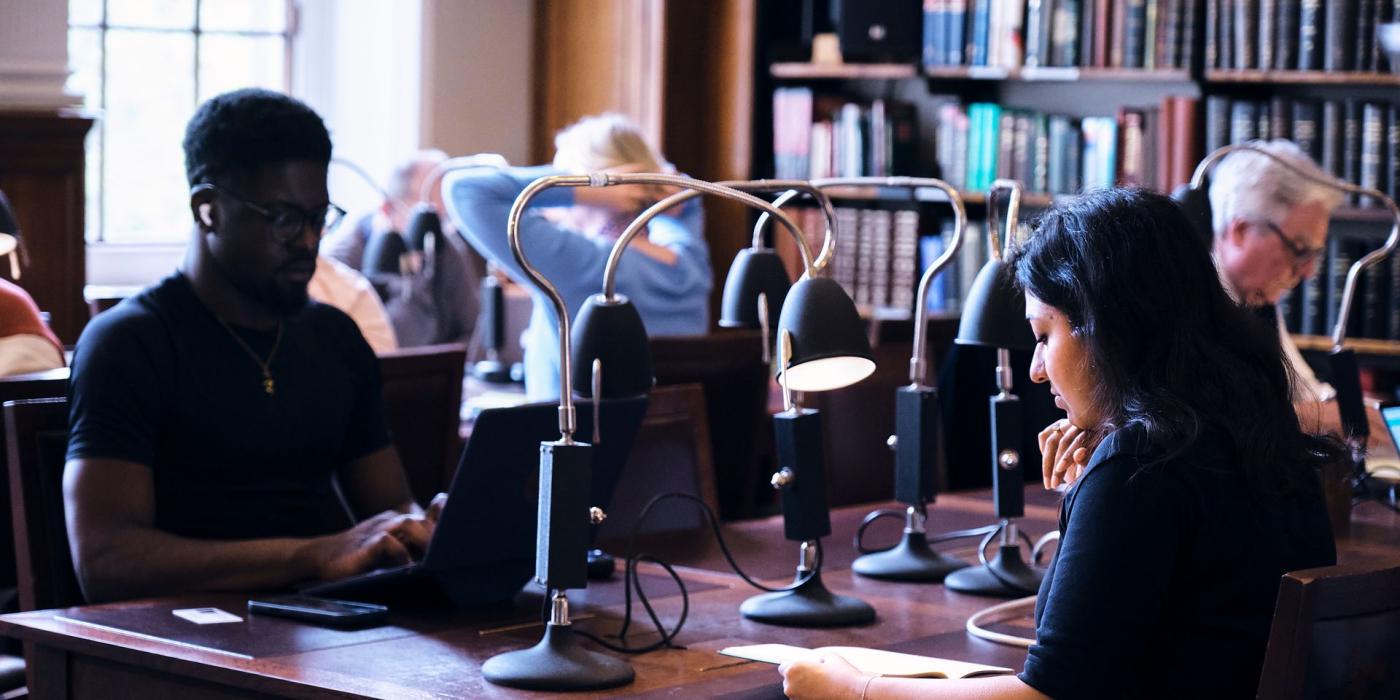 Readers working at a long desk beneath a row of angled reading lamps.