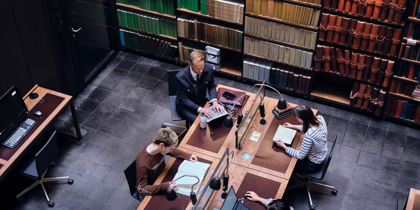 Overhead view of four members working at shared desks with reading lamps, beneath shelves of bound volumes.