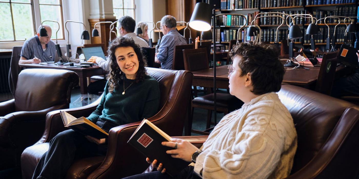 Two members talking in leather armchairs in the Reading Room, each holding an open book.