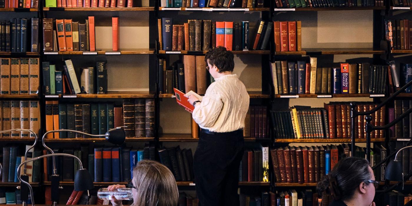 A member browsing floor-to-ceiling shelves with a red book in hand while others work at desks below.
