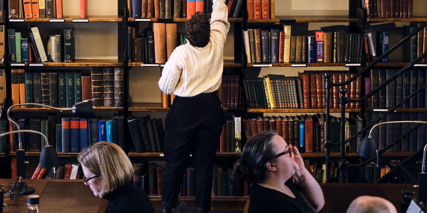 A member reaching for a volume on a high shelf as readers work at desks in the foreground.