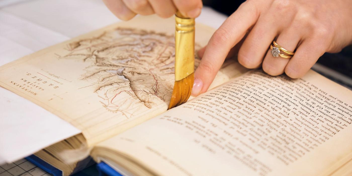 A conservator brushing paste along the gutter of an open book beside an engraved plate.
