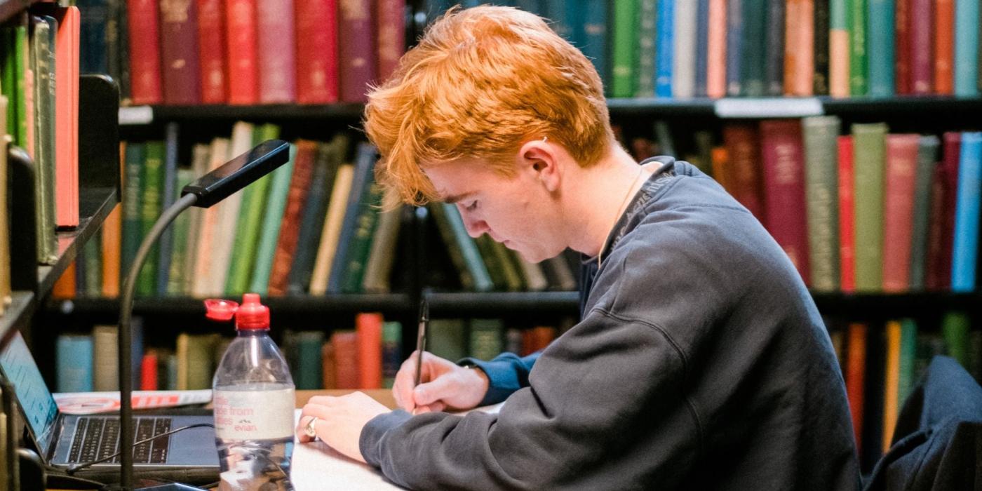 Young Man sitting at desk with book-shelf behind him.